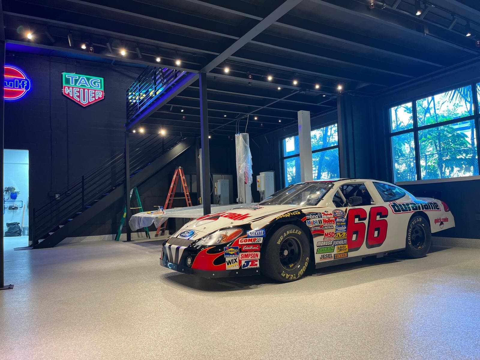 Finished clubhouse showroom with a liveried race car on a speckled epoxy floor, black steel mezzanine stair and neon signage